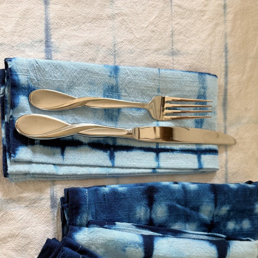 Indigo shibori napkins folded on a white cloth, with a fork and spoon pointing upward on a pale blue grid-patterned napkin