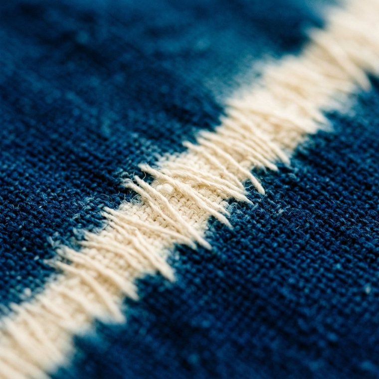 Extreme close-up of indigo-dyed cotton where deep blue meets a cream-white resist line, the dye feathering softly into the weave
